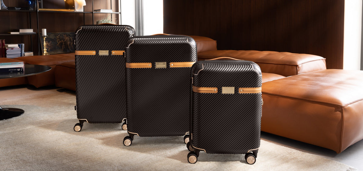 Three black hard-shell rolling suitcases with tan leather-like trim are arranged on a carpeted floor in a modern living room with brown leather seating and dark wood paneling.